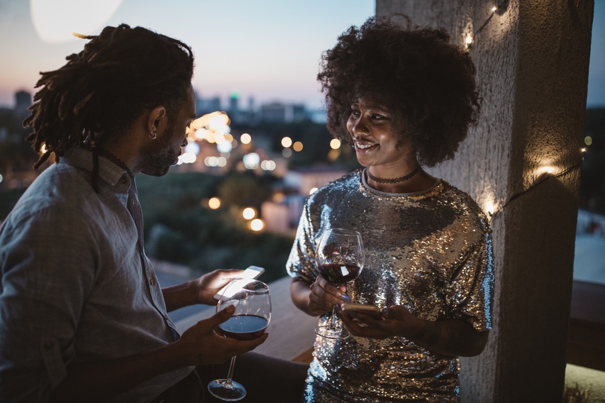 young couple having date night on balcony. Drinking wine, talking and surfing the net. Well dressed.