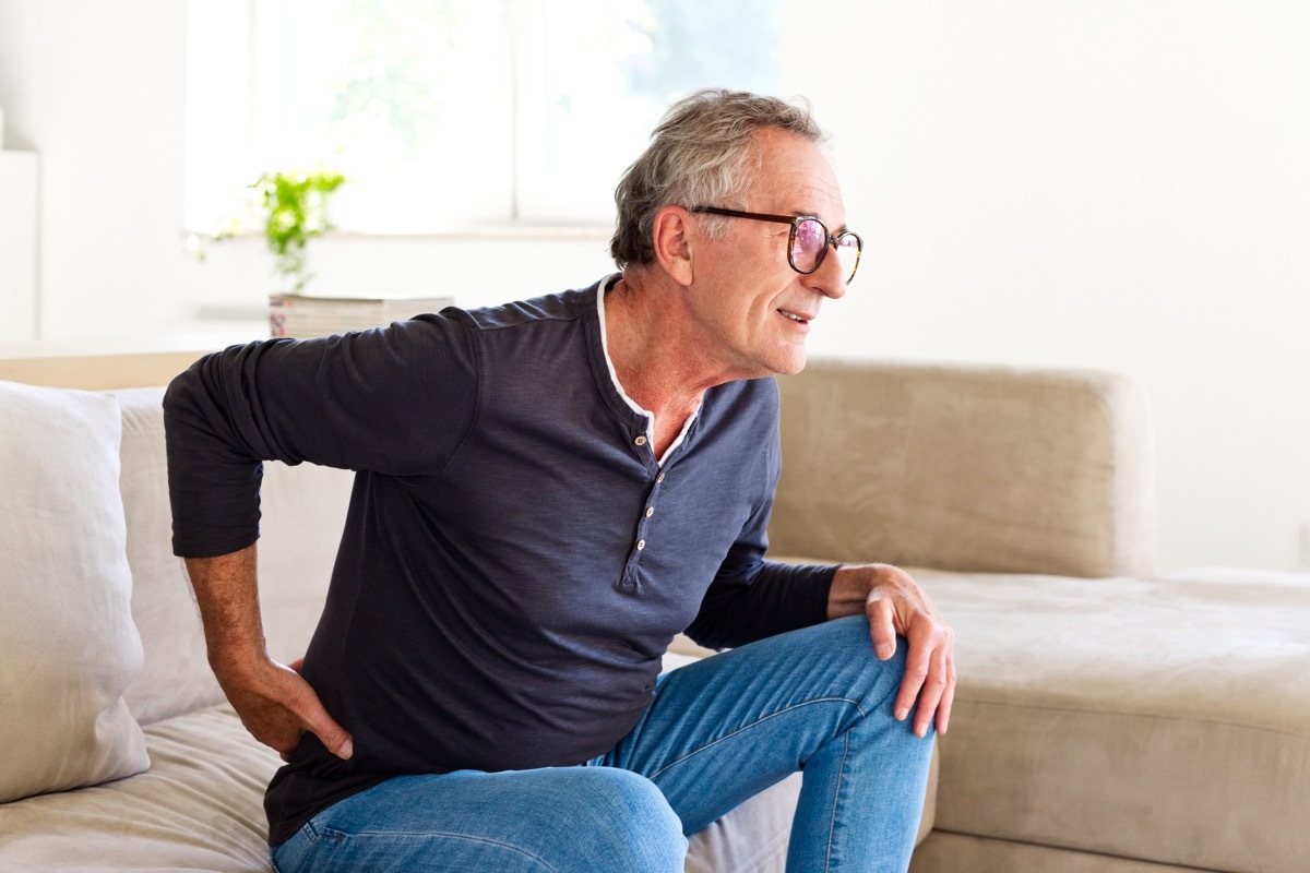 Worried senior man sitting on sofa at home and touching his back. Elderly man suffering lumbar spine.