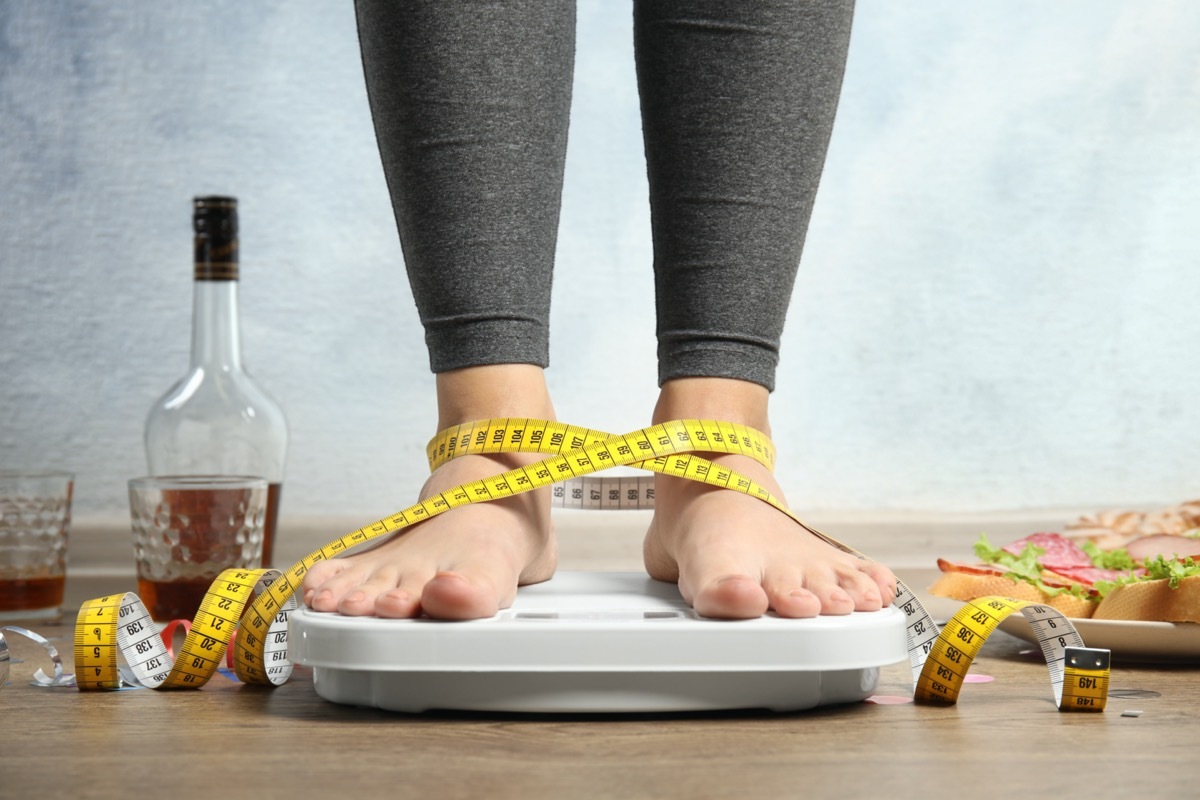 Woman with measuring tape using scale surrounded by food and alcohol after party on floor