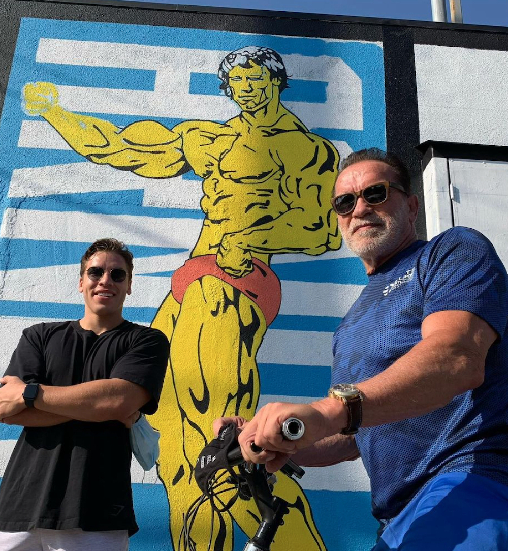 Joseph Baena and Arnold Schwarzenegger posing by a mural of Schwarzenegger