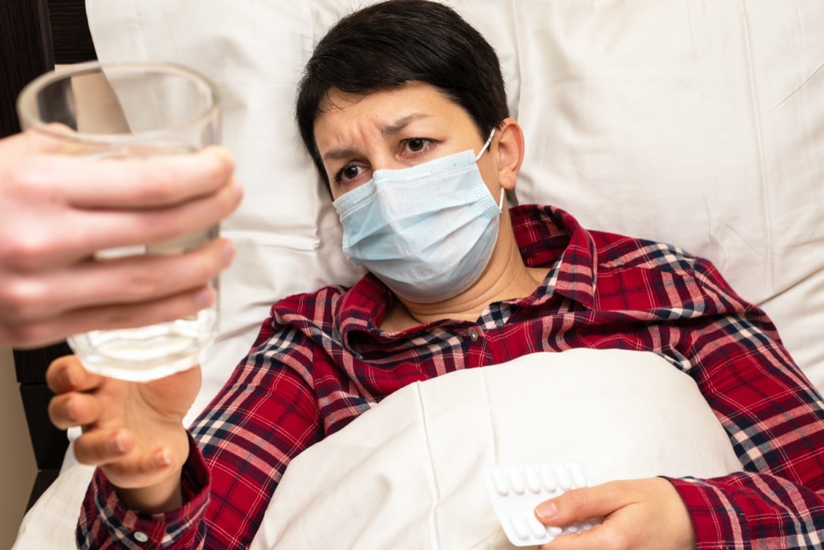 Sick woman in medical mask lying on the bed is given glass of water to take pills. Family care for the sick concept