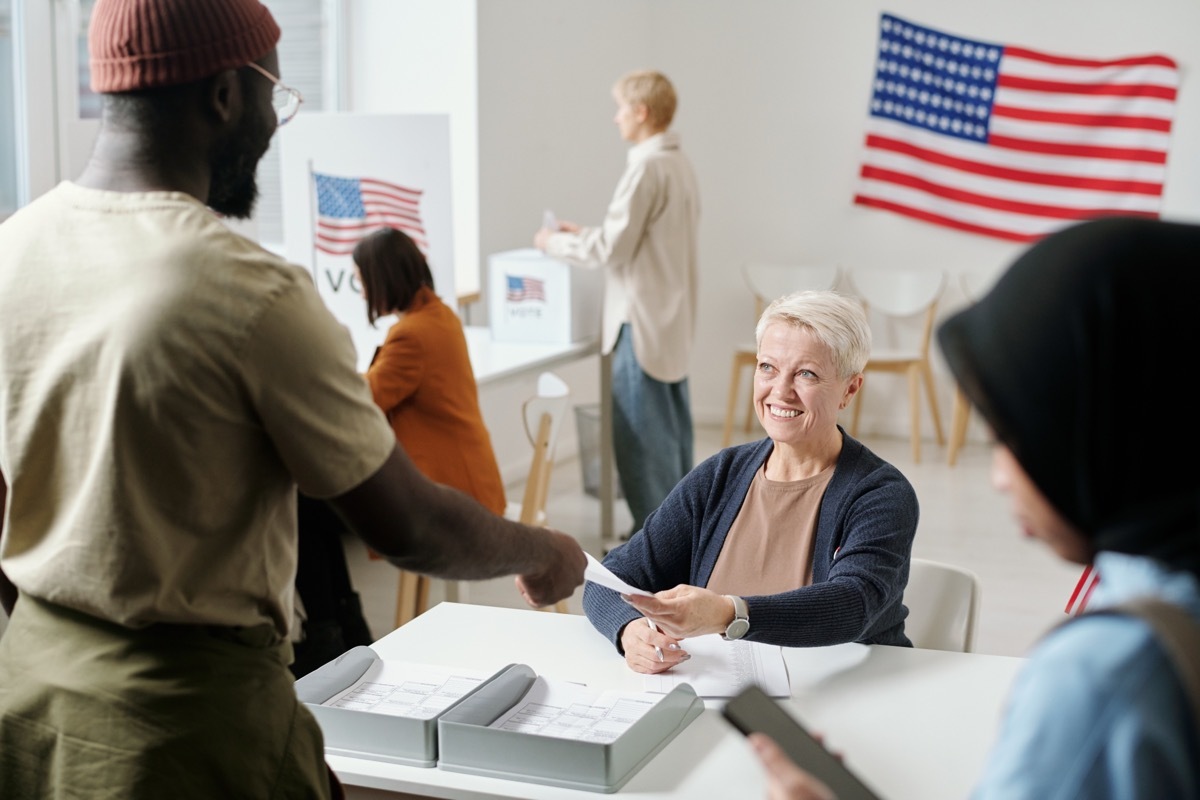 Happy blond mature woman passing ballot paper to young African American man standing by desk of electoral commission