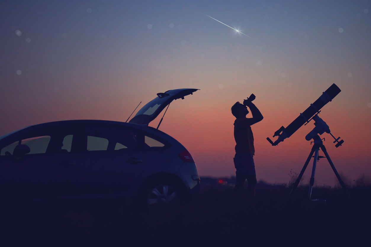 A silhouette of a man looking into the night sky with binoculars next to his car and a telescope while a meteor streaks overhead