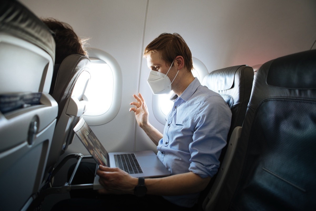 man in his 30s or 40s wearing face mask for COVID protection and waving at laptop in airplane seat