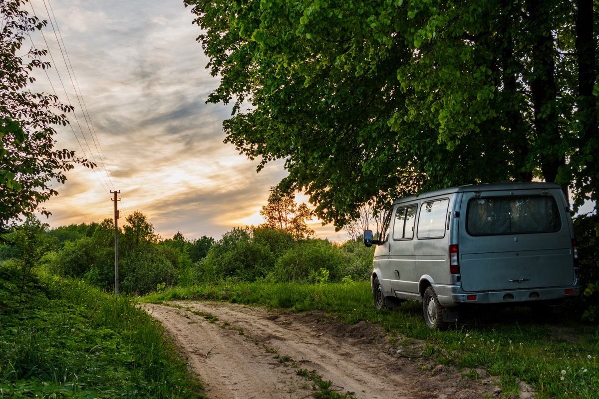 The minibus stands on the side of a dirt road. The machine stands under large linden trees. From behind you can see the sky and the sunset. - Image