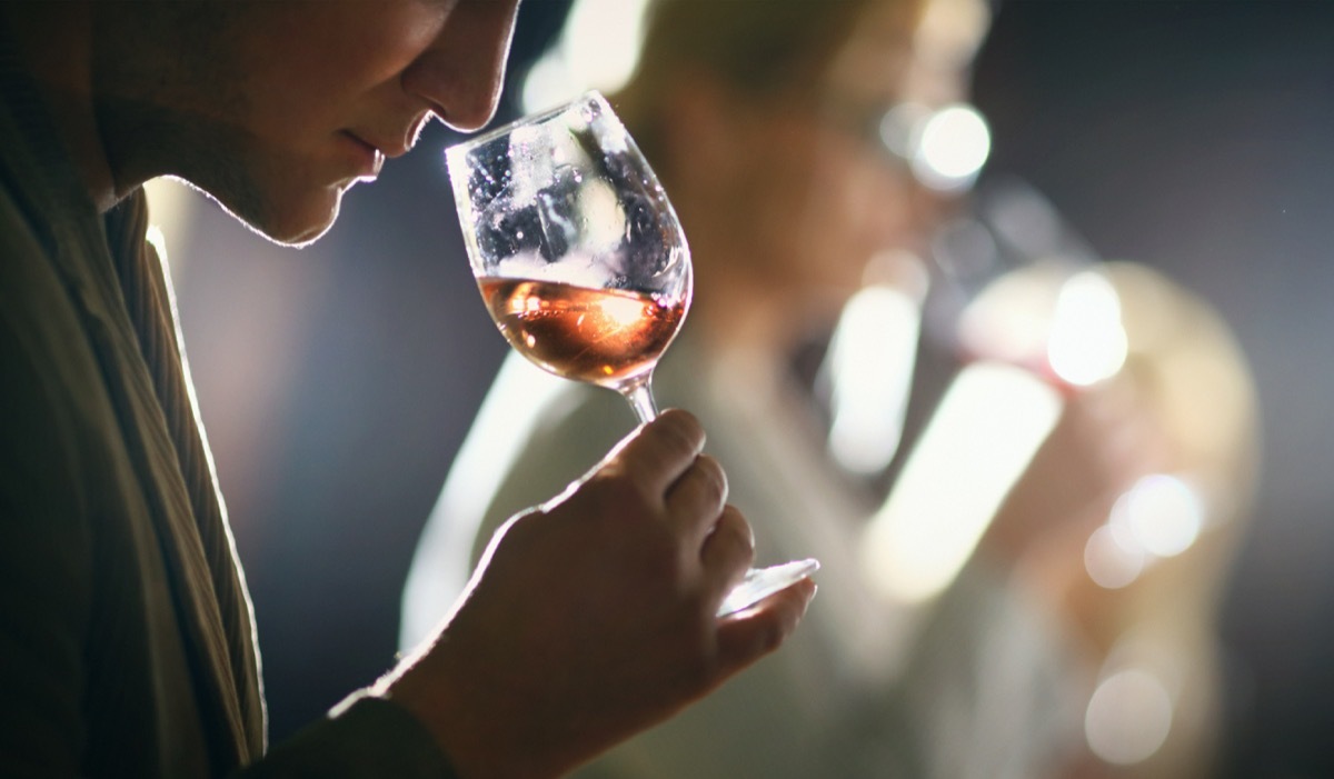 Group of unrecognizable caucasian adults tasting wine in wine cellar. Comparing appearance, smell, aroma,taste,aftertaste. The man in focus is holding a glass of rose wine and smelling the bouquet. There are two more people in background,blurry.