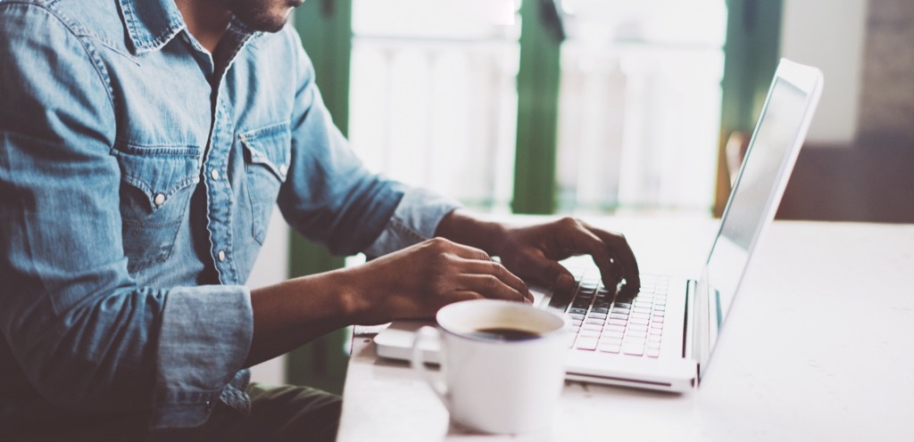 man in denim shirt types on laptop at desk with a coffee next to him, state fact about virginia