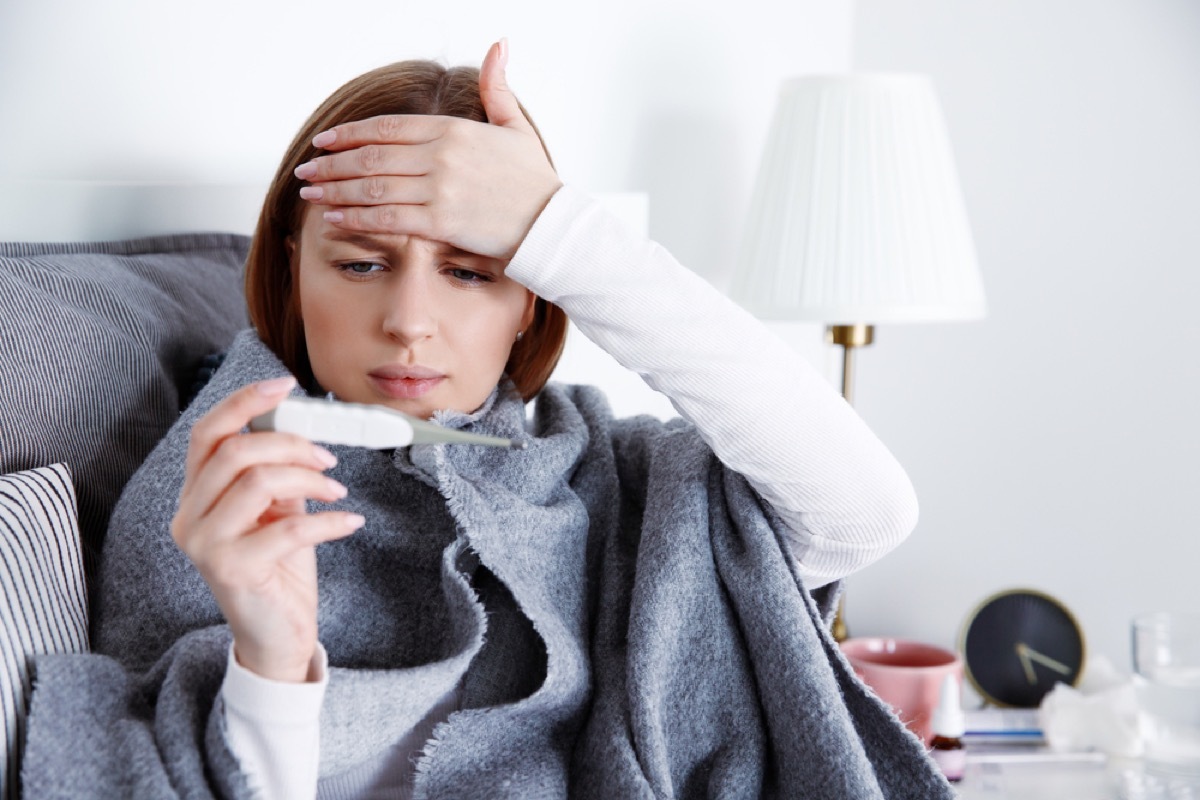 woman touching her forehead and reading a thermometer under a blanket