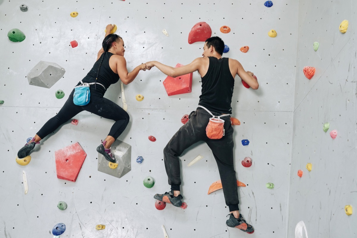 Back view man and woman working out in team and hitting hands while hanging on rock climbing wall