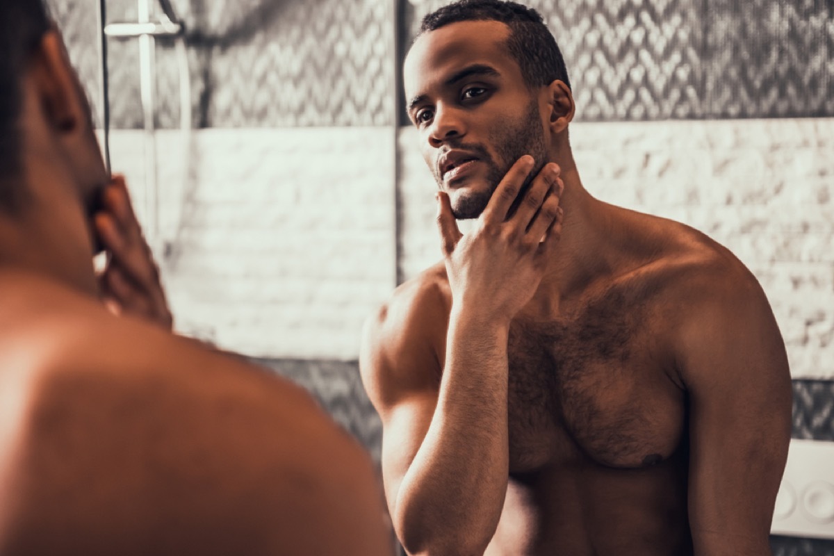 young shirtless man looking in mirror, moisturizing which helps fend off aging