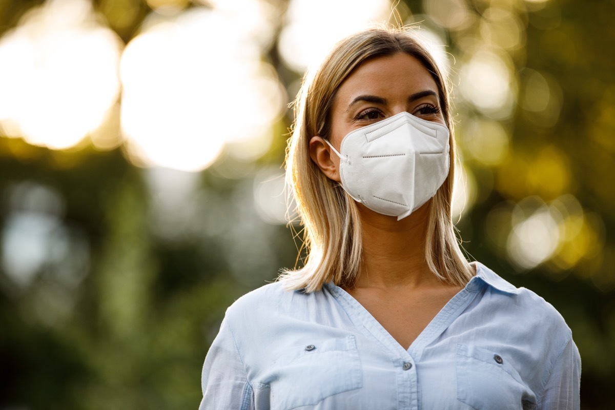 Copy space shot of happy young woman standing at the public park, enjoying a beautiful day out. She is smiling behind N95 face mask she is wearing and contemplating.