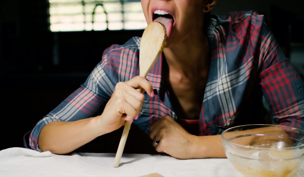 Woman licking batter off of wooden spoon in the kitchen