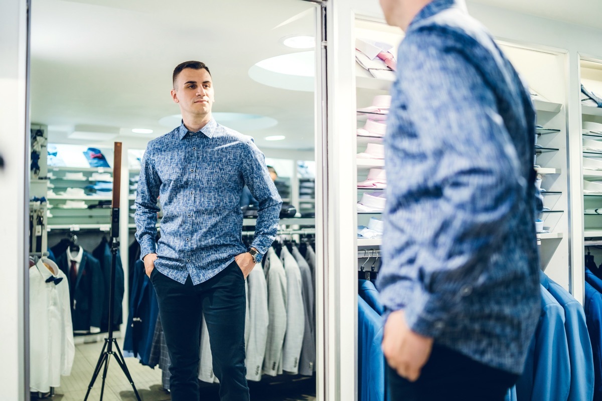 Young handsome caucasian man trying shirt in a store and looking himself in a mirror.