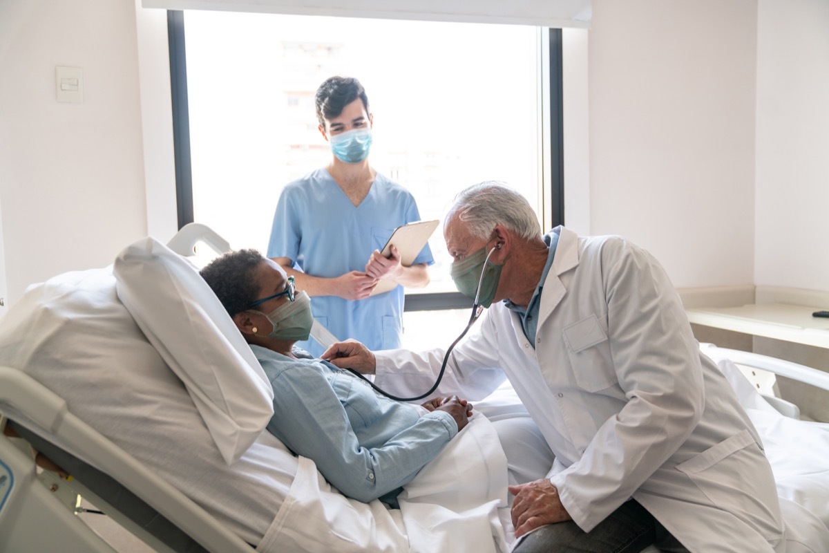 male doctor checking hospitalized female patient listening to her heart and nurse standing next to them all wearing protective facemasks - Pandemic lifestyles