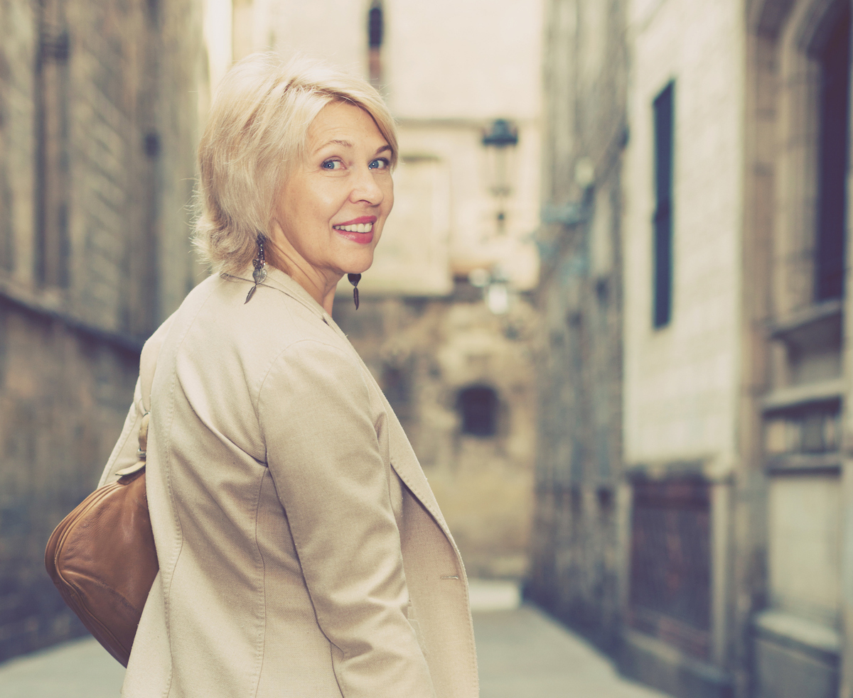 A smiling, mature woman with blonde hair and a beige jacket looks back towards the camera as she walks along a historic street