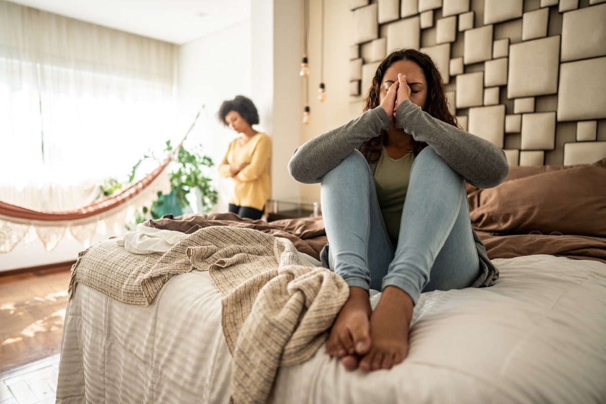 Woman sitting on bed upset with female partner looking on in the background