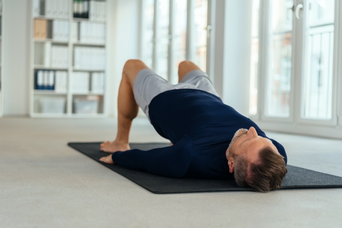 Man doing bridging exercise, lying on his back on black mat in empty office interior. Viewed from floor level from his head