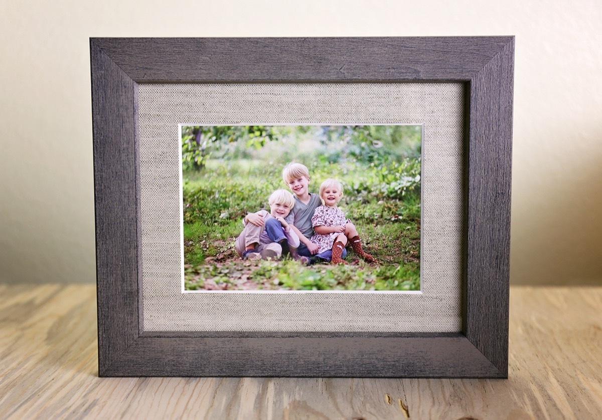 A tabletop display of a rustic wood framed print holding a portrait of a family of three happy young children sitting outside on a summer day.