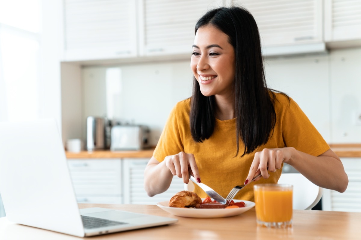 Image of a beautiful positive cheerful brunette young woman at the kitchen indoors at home using laptop computer have a dinner watch videos.