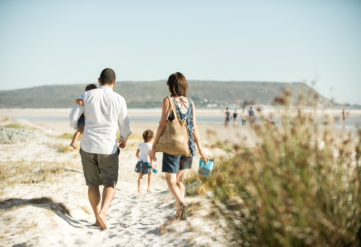 family walking on the beach outdoors in the sun, carrying their son while their daughter leads the way