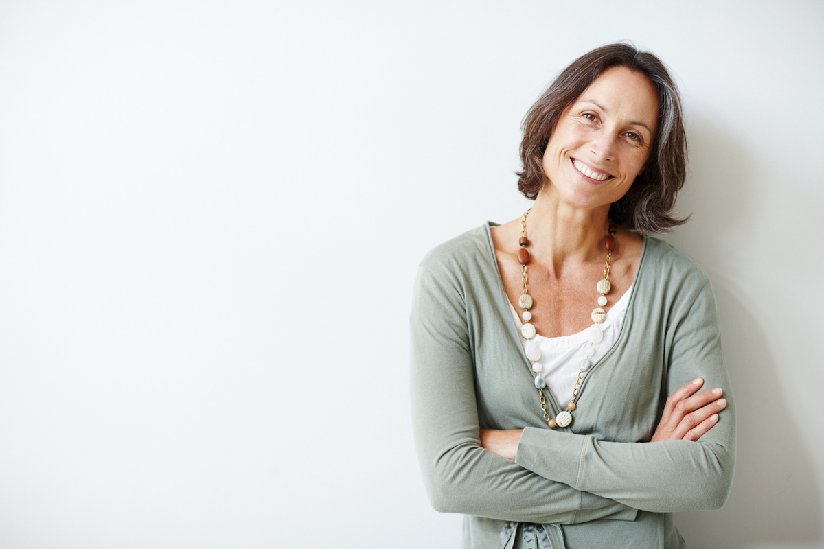 A smiling middle-aged woman with gray hair and a sage-green cardigan over a white t-shirt stands with her arms crossed against a pale gray background