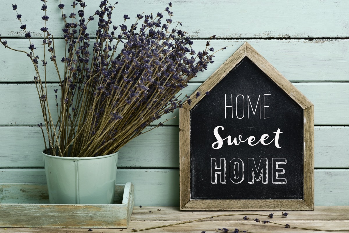 closeup of a house-shaped chalkboard with the text home sweet home written in it and a bunch of lavender flowers in a flower pot, against a rustic pale blue background