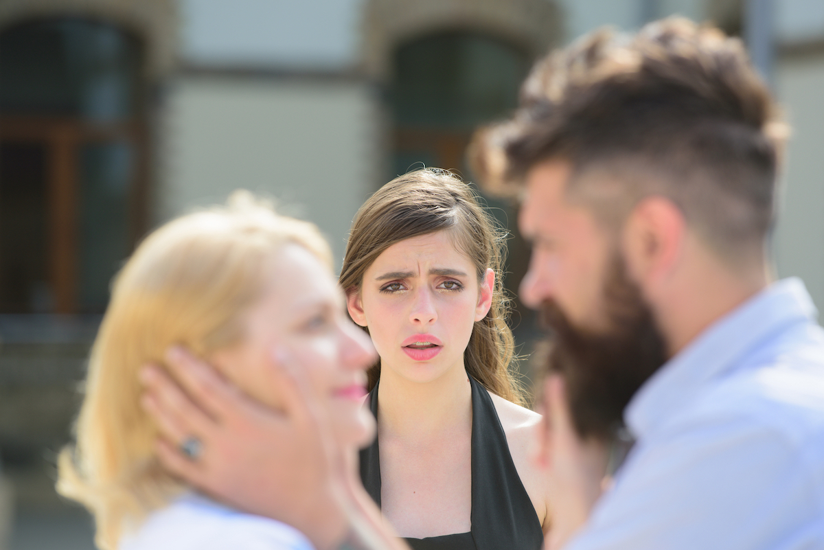 A young woman looking shocked and hurt behind her husband with another woman.