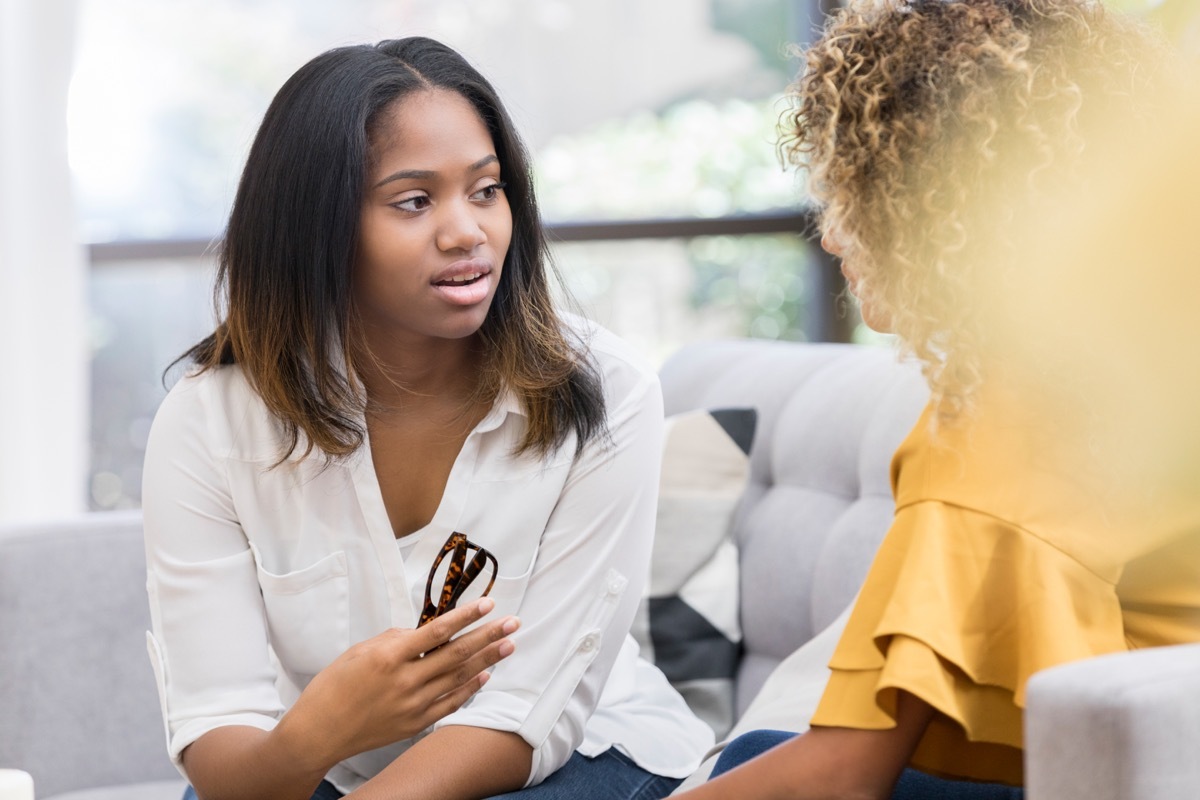 young woman is serious as she holds a pair of eyeglasses and sits on a living room couch with an unrecognizable friend. She is asking her advice on a matter of concern.