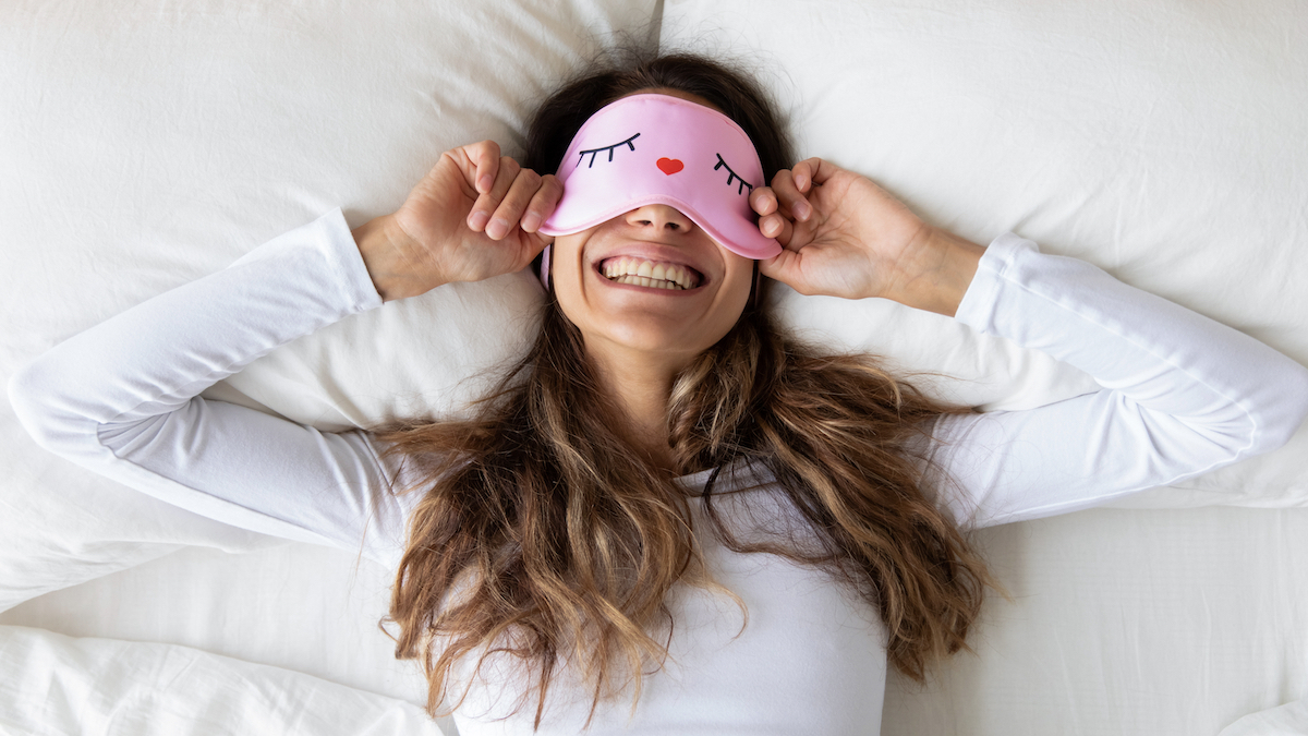 Top view of a happy woman wearing a funny pink sleeping mask enjoying morning lying in comfortable bed