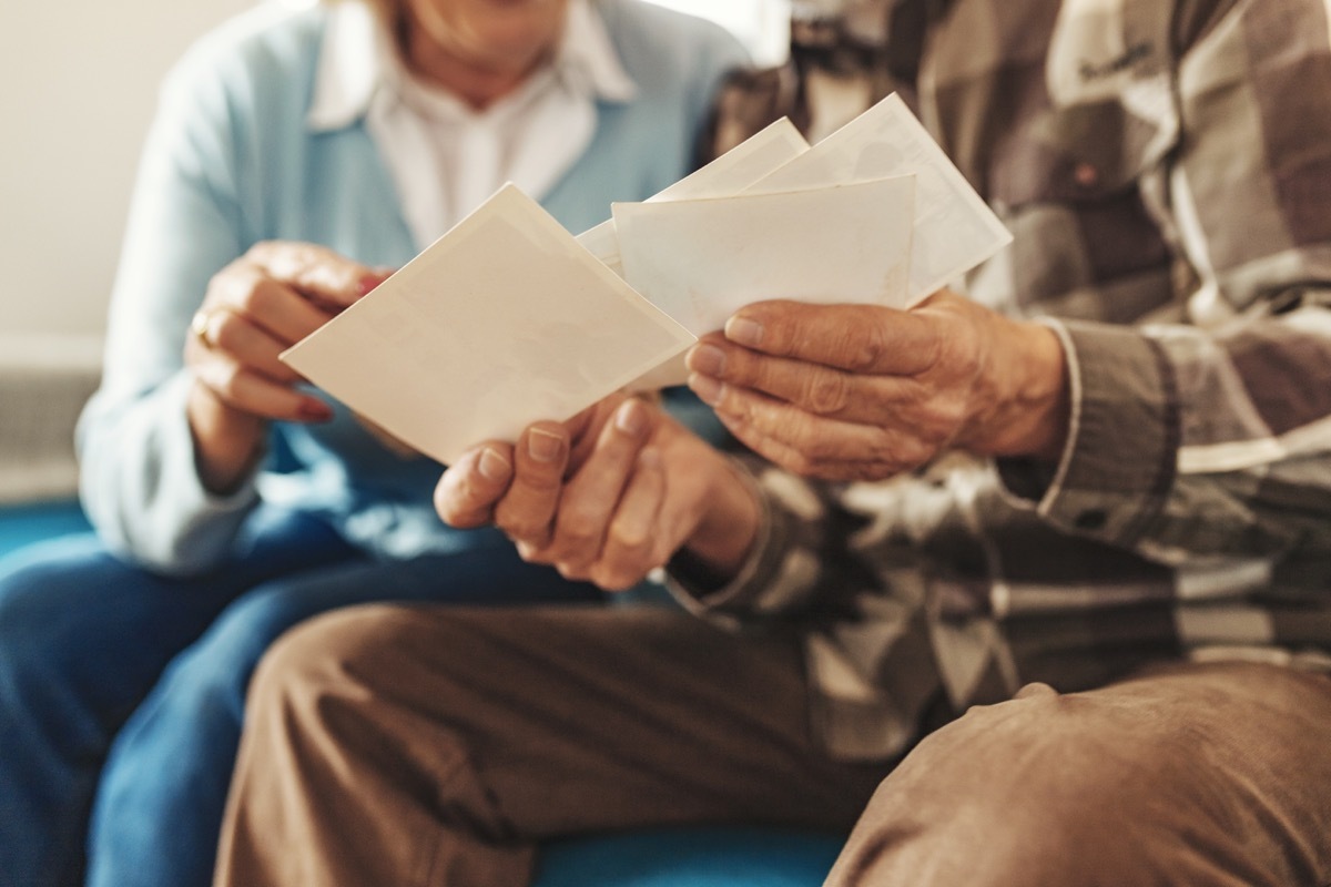  Cropped Image of Senior Couple Holding Up an Old Photos of Themselves When They Were Young While Sitting on Their Living Room. Concept of Active Playful Elderly During Retirement. Old People at Home Lifestyle