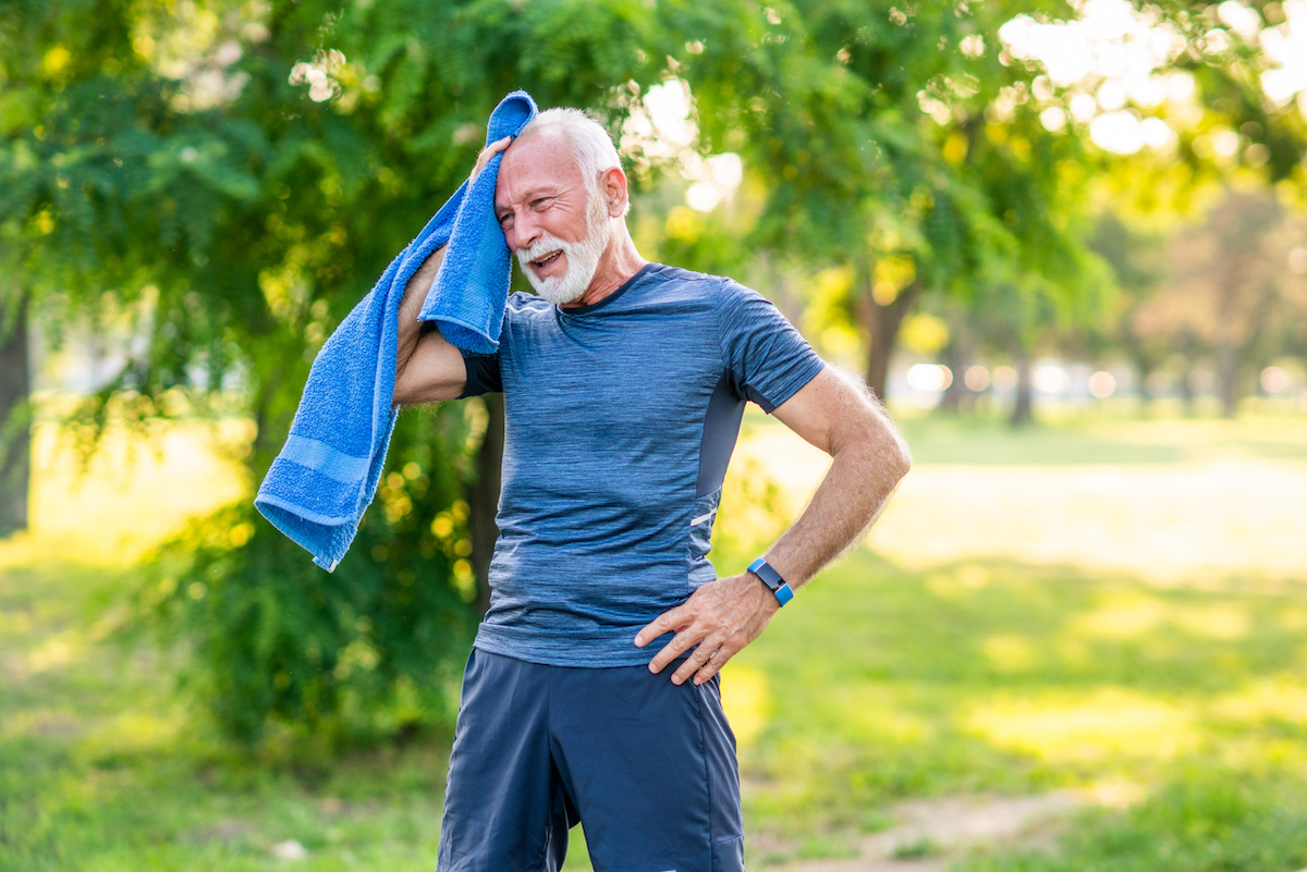 Senior man with white hair and beard wearing blue workout clothes takes a break while exercising outside on a hot day, wiping his face with a blue towel