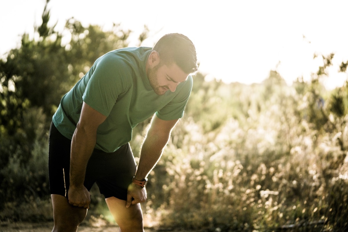 Tired male runner bending in forest. Mid adult jogger is in sports clothing. He is exercising on sunny day in woods.