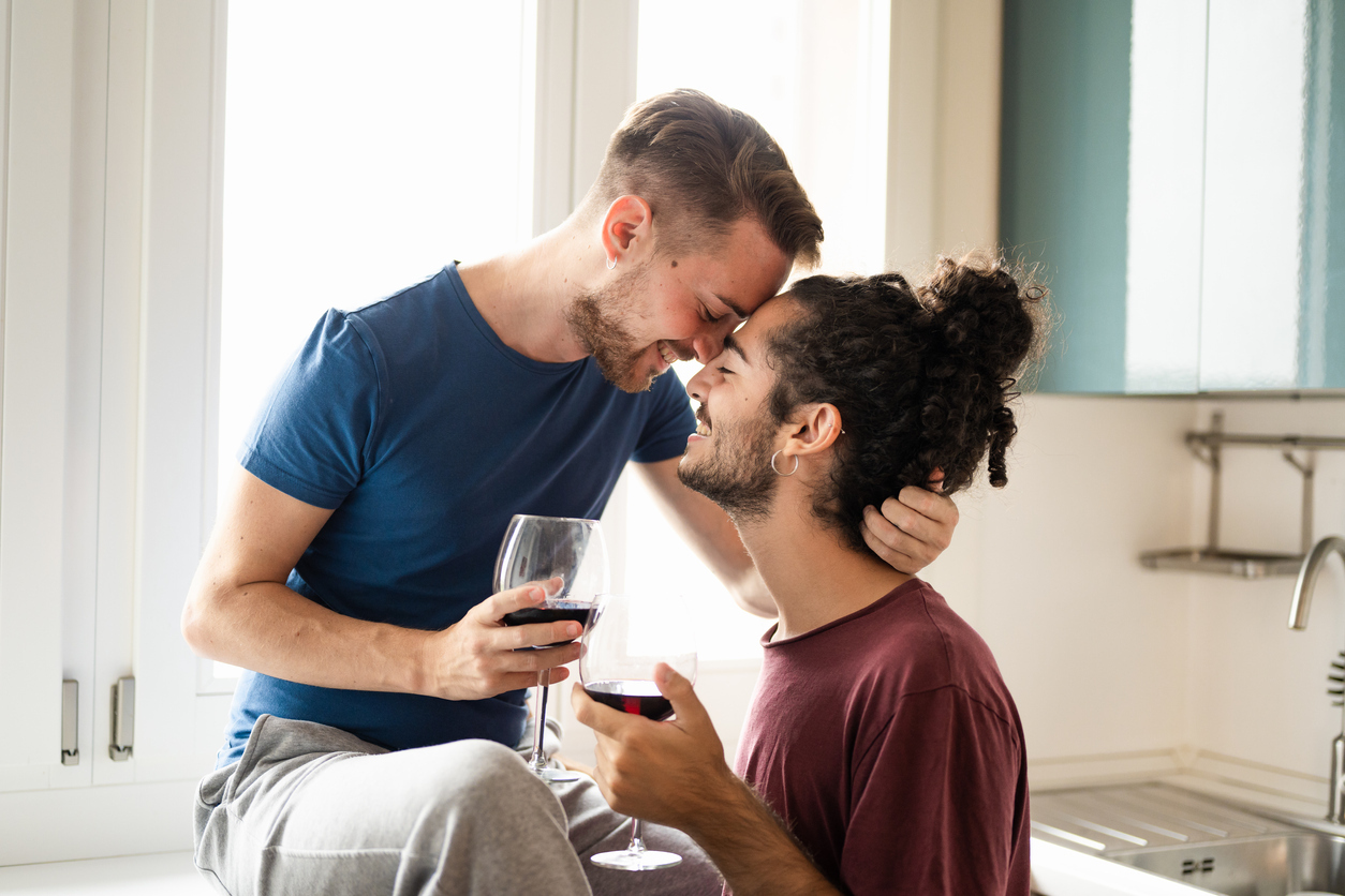 Tenderness moment of smiling gay couple, young men toasting with red wine in the kitchen, male couple having fun and drinking red wine for celebration of their engagement