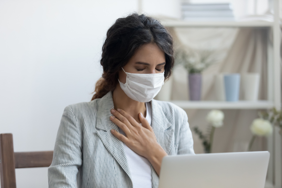 Business woman in mask sitting at desk is suffering from repeated coughing and breathing difficulties