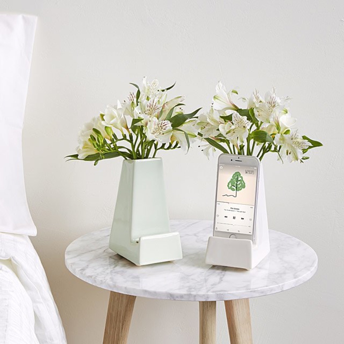 two ceramic vases with flowers and a smartphone on a marble topped nightstand next to a bed