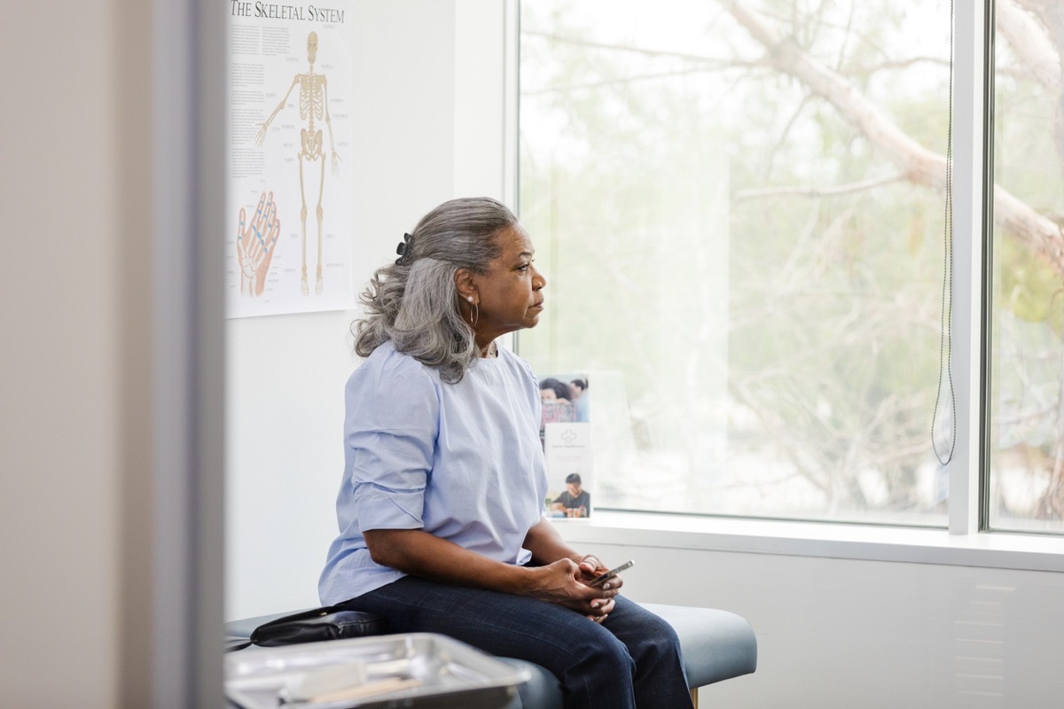 mature woman sits on the examination table and holds her mobile phone as she waits for her doctor to enter the room.