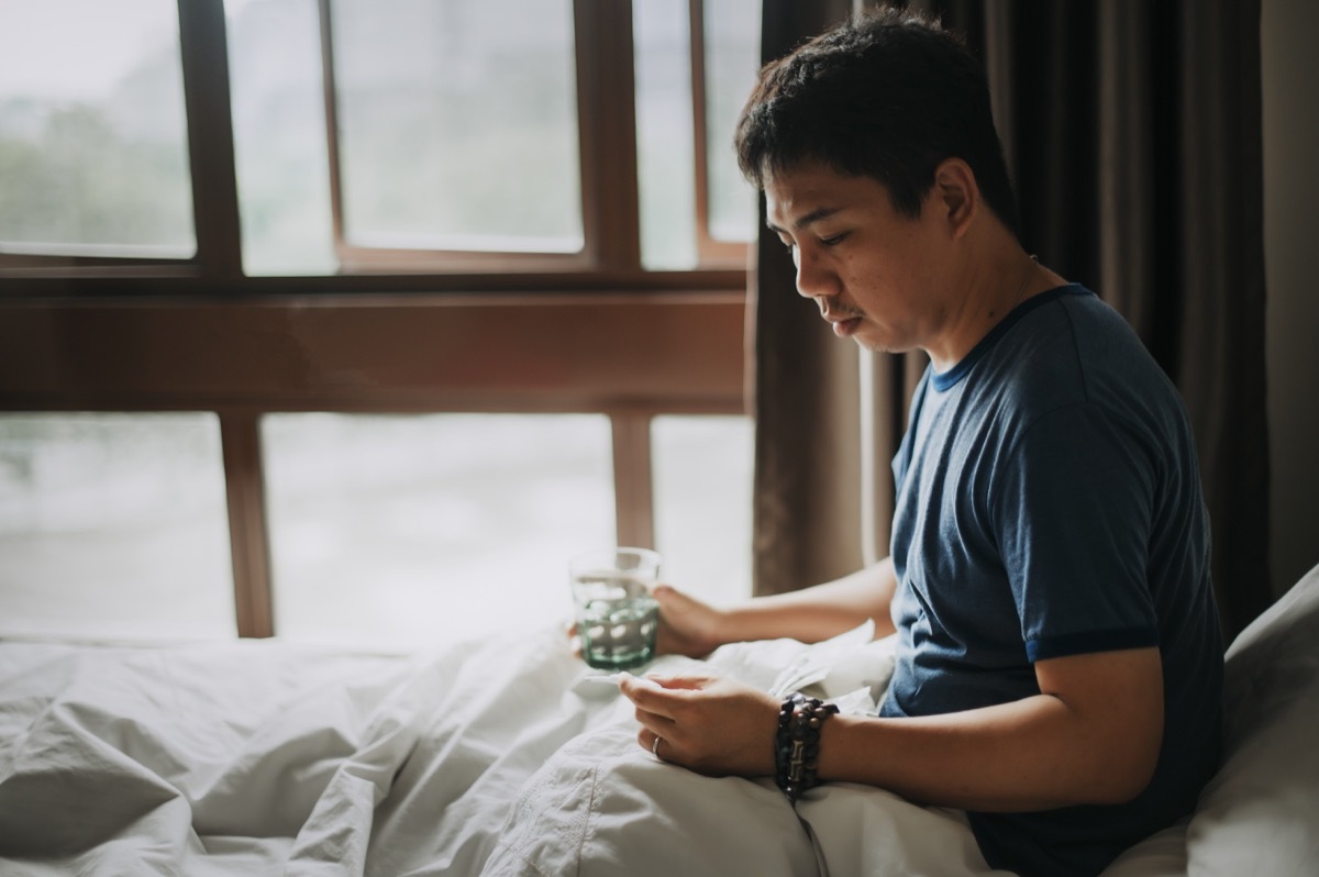 male sick illness at home and checking his body temperature using a thermometer and taking medicine with water on a bed